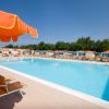 Piscina vuota con lettini e ombrelloni arancioni, cielo sereno e alberi sullo sfondo.